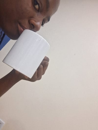 The scene is located in the city of Close, Nigeria. A dark-skinned individual, gender indeterminate, is visible in the left portion of the frame. Their lower face, including the nose and pursed lips, is in direct contact with the rim of a plain white, cylindrical mug. The individual's right hand grips the mug. A blue garment is visible on the upper left, possibly part of a collar or uniform. The background consists of a uniform, light-colored wall with minor textural variations, indicating an indoor environment. The individual is engaged in the act of drinking from the mug.