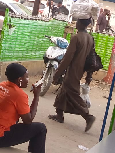 The image captures an outdoor street or market scene during what appears to be daytime. The setting suggests a lively, possibly urban or semi-urban environment, with elements of commerce and daily life.
In the immediate foreground on the left, a woman is seated, mostly seen from behind but slightly turned to her right. She is wearing an orange short-sleeved top with white text visible on her back, partially reading "TTALL" and "ll Kmbwe". She has a black head covering and dark trousers. She is actively engaged with a white smartphone held to her left ear, and distinctive white, elongated earbuds are clearly visible in both of her ears, suggesting she is on a call or listening to audio.
In the midground, moving from right to left, a man is walking away from the viewer, seen from the back and side. He wears a long, loose-fitting dark brown garment, likely a traditional tunic, over dark trousers and dark athletic shoes. A dark head covering is also on his head. He demonstrates a common practice of carrying goods on his head, balancing a large, irregularly shaped white bundle. Additionally, he carries a black bag slung over his left shoulder and a smaller white plastic bag in his right hand.
Behind these two individuals, the scene unfolds further. A white and light blue scooter is parked between them. In the background, a green plastic barrier or banner stretches across the scene. This barrier is covered with repetitive white text, which, according to OCR, includes variations of "oromo", such as "oralmo", "oramo", "orakIO", "Bromo", "moyomo", "pocromo", "onoma", and "orcima". Behind this green barrier, several parked cars, mostly light-colored, are visible. Other people are also present in the background, including a man in a brown outfit on the far right holding a phone to his ear, and more indistinct figures further back near the cars. The ground appears to be a mix of dirt and possibly some paved sections. A blue and green striped pole or pillar is visible on the far right edge of the frame.
The overall activity depicted is people going about their daily routines, with the woman on the phone and the man transporting goods, contributing to a sense of a bustling street or market atmosphere.