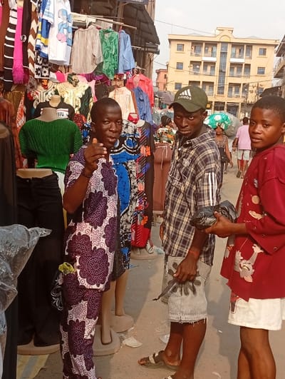 The image depicts a street market scene in Nkwelle, Nigeria. In the foreground, three males are present:
1. On the left, an adult male wears a purple and white patterned long-sleeved shirt and trousers. He faces the viewer, extends his right arm, and points with his index finger. A small green object is visible in his left trouser pocket.
2. In the center, a young adult male wears a green Adidas baseball cap, a blue and white plaid short-sleeved shirt, ripped light-wash denim shorts with a black starburst design, and dark sandals. He holds a dark, irregular-shaped object in his left hand and looks directly towards the viewer.
3. On the right, a young male wears a red short-sleeved shirt with a cartoon character print and white shorts. He holds a black plastic bag with both hands and looks towards the viewer.
Behind the foreground individuals, numerous articles of clothing are displayed on racks and mannequins, including blouses, dresses, and shirts of various colors and patterns, indicating a clothing stall. In the midground, several pedestrians are visible, including a woman carrying a large bundle on her head and a man in a pink shirt. The background features a multi-story building with a light-colored facade and numerous windows, suggesting urban infrastructure. The ground surface appears paved with some scattered debris. A partially visible sign overhead indicates "MARKET STREET."