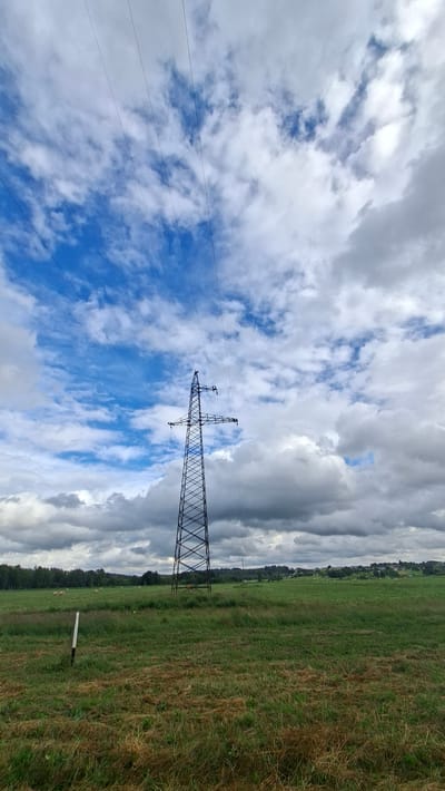 The image shows a wide-angle, low-angle view of an open, rural landscape under a dramatic sky. In the center of the frame, a tall, lattice-like electricity pylon stands prominently, its metal structure stark against the sky and receding into the distance. Multiple power lines extend from the top of the pylon and disappear towards the upper left corner of the image. The sky is a mix of bright blue patches and heavy, white and grey clouds, suggesting dynamic weather conditions. The foreground and midground are occupied by a large, grassy field with varying shades of green and brown, indicative of mown or drying grass. A white and black striped pole is visible in the left foreground. In the distance, a treeline marks the horizon, with hints of a village or scattered buildings beyond it. The scene appears to be located in a rural area, possibly agricultural land.