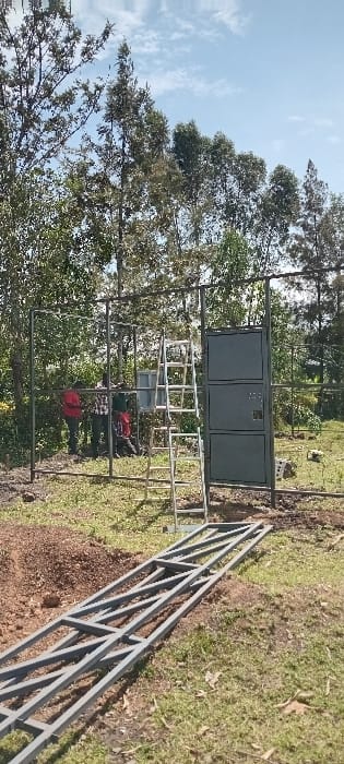 The image displays an outdoor construction scene in Kisumu, Kenya, under a partly cloudy sky. Three adult males are observed in the midground, engaged in the assembly of a dark gray metal frame structure. The male on the far left wears a red shirt, the central male wears a plaid shirt, and the male to the right wears a red shirt; all appear to be manipulating or attaching components to the frame. A partially erected dark gray metal fence or enclosure structure stands upright, with a three-paneled metal gate section installed on its right side. A silver-colored A-frame stepladder is positioned upright in front of this structure. In the foreground, two long, dark gray metal truss-like sections are laid horizontally on the ground. The ground cover consists of sparse green grass intermixed with piles of reddish-brown soil. A dense treeline of tall, slender trees forms the background.