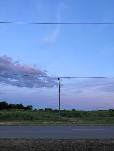 The image shows a road running alongside a grassy field with trees in the distance. A power line stretches across the frame, supported by a utility pole with a street lamp. The sky is a soft blue with hints of pink and purple, suggesting it is either sunrise or sunset. The overall mood is peaceful and serene. The image is oriented vertically, with the road and field on the right side and the sky on the left.