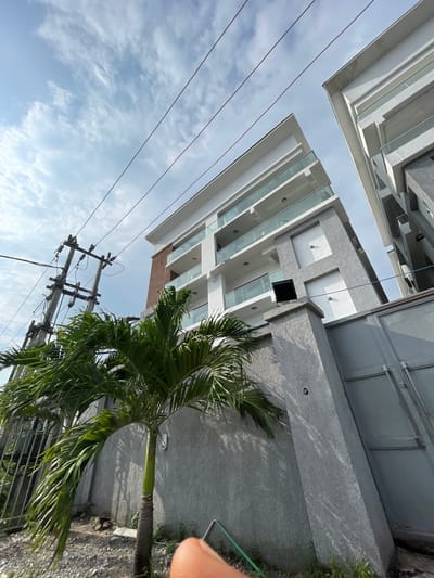 The image shows a multi-story building with balconies and glass railings, located in Ilado, Nigeria. A palm tree is in the foreground, and power lines are visible against a cloudy sky. The building appears modern, with a combination of white and grey facade. The overall impression is an urban landscape with a touch of tropical vegetation.