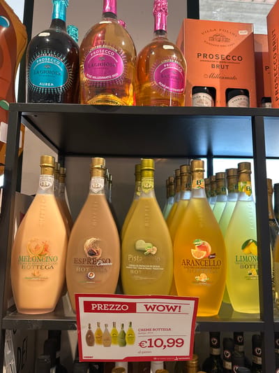 The image shows a shelf in a shop displaying various bottles of alcohol. On the top shelf, there are several bottles of Prosecco, including "LAGIOSA" in both blue and pink labels, and a boxed set of "VILLA FOLINI" Prosecco Brut. Below, on a lower shelf, a row of Bottega liqueur bottles is visible. These include "Meloncino," "Espresso Cream," "Pistacchio," "Arancello," and "Limoncello." A red and white sign is placed in front of these bottles, displaying "PREZZO" and "WOW!" with a price of "€10,99" for "CREME BOTTEGA." The bottles are arranged neatly on black shelves, and the background appears to be a retail environment, possibly a supermarket or liquor store. The image is taken from a slightly elevated angle, looking down at the products. Based on the presence of Italian brands and products like Prosecco and Bottega liqueurs, this scene is likely located in Italy. The specific mention of "Tolentino, Italy" in the prompt is not directly verifiable from the visual information in the image alone, but the context of Italian alcohol brands strongly suggests a location within Italy.