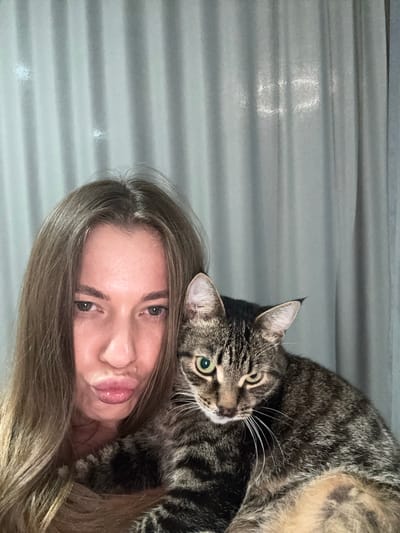 The image is a selfie taken by a young woman with long, blonde hair, who is lying down. A tabby cat is lying on top of her, with its head resting on her face. The cat has green eyes and is looking at the camera. The woman is smiling and appears to be in good spirits. The background is a textured gray surface, possibly a wall or a blanket. The lighting suggests it might be daytime, but the specific time of day or weather conditions are not discernible. The setting is intimate and personal, indicating a home environment. There is no visible text in the image. The woman's hair is spread out around her face, and the cat's fur is detailed and striped. The image conveys a sense of affection and companionship between the woman and her pet.