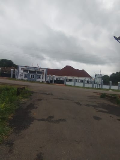 The image shows the Ambassador Hotel in Ogbomosho, Nigeria, under a cloudy sky. The hotel has a white facade with gray accents and a red-tiled roof. Flags are visible flying above the entrance. A paved road leads to the hotel, with grass and bushes on either side. In the foreground, there are patches of water on the road. A street lamp is visible on the right side of the image.