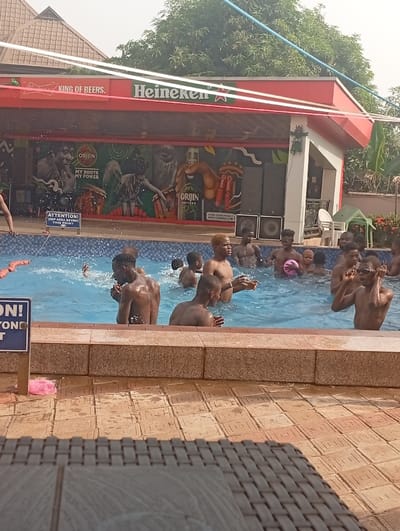 A daytime scene shows a crowded swimming pool with numerous people of various ages enjoying the water. Behind the pool is a bar structure with "KING OF BEERS" and "Heineken" signs atop. A mural featuring Orjin products decorates the back wall. A warning sign reads "ATTENTION! KEEP AREA BEYOND THIS POINT." The foreground reveals a patio with tiled flooring and a wicker table. The overall ambiance suggests a lively and recreational environment.