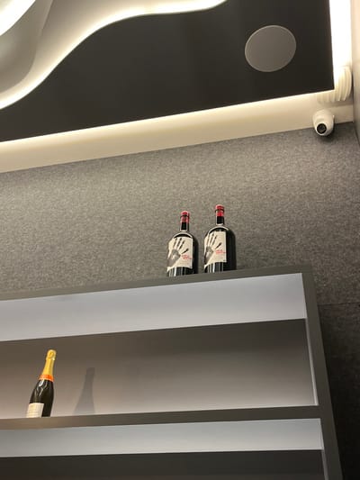 Two bottles of red wine, labelled "Mano Garcia" are on the top shelf of a white shelf unit with a grey, textured background. A single bottle of champagne sits on the lower shelf. The shelf unit appears to be in a bar or restaurant in the city of Las Palmas de Gran Canaria, Spain, at the coordinates 28.102, -15.414. The top of the shelf is illuminated by a curved LED light fixture that appears to be part of a larger ceiling fixture. Above the light fixture is a white ceiling panel and a circular white speaker. A white security camera is mounted on the wall near the ceiling, slightly offset from the ceiling light.