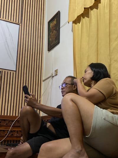 This image captures an intimate moment between two people in a domestic setting in Sukasada, Indonesia. The primary subjects are a man and a woman. The woman, with dark hair, leans over the man, her face close to his. She is wearing a brown top and light-colored shorts. The man, wearing glasses, is looking down at his phone, which he holds in his right hand. He is dressed in a black t-shirt and black shorts.
The background features a textured, golden curtain or drapery, suggesting an indoor environment. Below this, a wall with horizontal wooden slats is visible, a common design element in some Indonesian homes. A framed picture hangs on the wall above the wooden slats, depicting what appears to be a scene with figures. Electrical outlets and wiring are also visible on the ceiling area. The lighting suggests it is likely evening or indoors with artificial lighting.
The overall mood seems to be relaxed and personal, with the woman's gesture indicating affection or concern towards the man engrossed in his phone. The scene implies a private moment within a home. No visible text is present in the image.