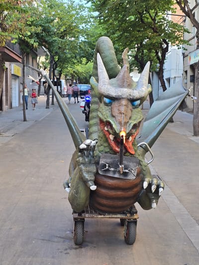 This image captures a close-up view of a dragon float being prepared for an outdoor event, likely a parade or festival, given the presence of onlookers and a street setting in Cornellà de Llobregat, Spain. The dragon, a large, green, and brown structure, appears to be mounted on a wheeled cart for mobility. Its head is detailed with menacing horns, blue eyes, and open jaws, from which a jet of fire is emanating, suggesting an active pyrotechnic element of the display. Several metal clips and supports are visible, indicating ongoing setup or maintenance.

In the background, trees with lush green foliage line a paved street. A few people are visible, some standing and observing, while others appear to be participating or working on the float. A car with flashing blue lights, possibly a police vehicle, is parked on the street. The lighting suggests it is daytime, with natural light illuminating the scene. The overall atmosphere seems to be one of anticipation and preparation for a public celebration. No visible text is present in the frame.