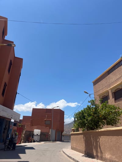 The image, taken in Ouaouizert, Morocco, shows a street scene under a bright blue sky with scattered clouds. Buildings line the street, constructed in earth-toned hues. Power lines stretch across the sky between the structures. A tree with green foliage and what appear to be citrus fruits grows near the top right building. A partially visible mountain can be seen in the distance.

On the right side of the street, a person in a jacket and helmet stands near a parked motorcycle. A stack of rectangular containers and construction materials are present nearby. A sign with lettering is partly visible near the bottom of the image. The street pavement shows visible wear and tear.