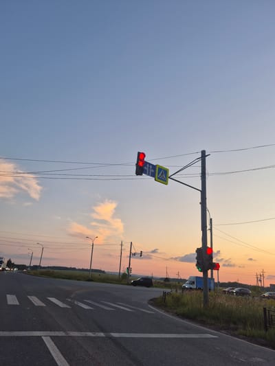 The image shows a road intersection at dusk in Novoe Mart'yanovo, Russia. A traffic light with a red signal is visible, along with a pedestrian crossing sign. Several cars are present on the road, including a white van and a black SUV. The sky is a gradient of blue and orange, with some clouds. The environment appears to be rural or semi-rural, with fields and trees visible in the background. There are no people clearly visible in the image. Power lines are strung across the scene.