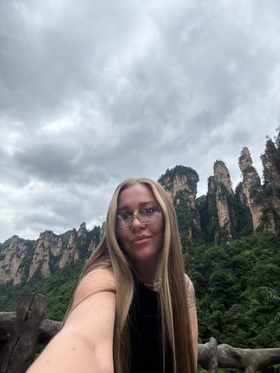 A person with long, blonde hair and glasses is taking a selfie against the backdrop of the Zhangjiajie National Forest Park in Luoguta, China. The towering sandstone pillars characteristic of the park are visible behind her, partially obscured by lush green vegetation. The sky is overcast and cloudy. The person is wearing a black top. A wooden railing is visible in the foreground. A tattoo is visible on the person's upper arm.