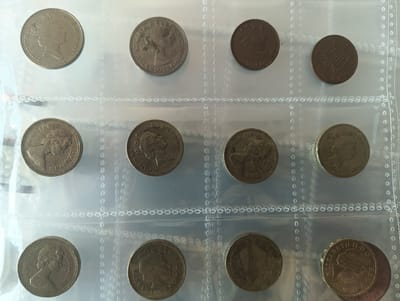 The image displays a collection of twelve coins, each encased in individual plastic sleeves within a clear, protective binder sheet. The coins vary in color, size, and design, suggesting different denominations and origins, possibly British. Several coins show portraits of British monarchs, including Queen Elizabeth II, and other monarchs. Some coins have visible dates, such as 1944, 1977, 1983, 1984, 2006, and 2010, and inscriptions like "Elizabeth II D G REG F D" are visible on some of the coins. The plastic sheet has a series of holes along its borders, indicating it can be placed in a ring binder. The image appears to be taken indoors under natural lighting. There is no information to indicate that the image was taken in Karnobat, Bulgaria.