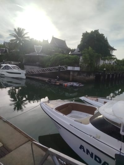This image depicts a marina in Ko Kaeo, Thailand, likely within a resort or private residence. The scene is characterized by tranquil waters reflecting the overcast sky and lush tropical vegetation. In the foreground, the stern of a white motor yacht with brown upholstery is prominently featured, displaying the partial name "ANDA" on its hull. Another white motor yacht is docked to the left, with its registration number "DB 37" visible. A gangway connects the yachts to a stone-laden dock, behind which are several buildings with distinctive pitched roofs, reminiscent of traditional Thai architecture, surrounded by dense greenery and palm trees. A few jet skis are also visible in the water. The overall impression is one of a peaceful, upscale waterside environment.