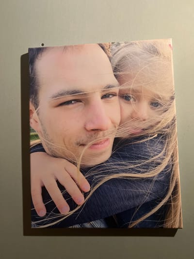 The image is a close-up photograph of two people, a man and a child, seemingly embracing. The man's face is in the foreground, slightly out of focus, with his eyes closed and a gentle smile. The child, with blonde hair, is nuzzling against the man's face, their eyes looking directly at the camera. Both subjects have fair skin. The man appears to be wearing a dark blue shirt, and the child's blonde hair is falling across both of their faces. The overall impression is one of affection and closeness. The lighting suggests it might be indoors or during daylight hours, but the precise time of day or weather conditions are not discernible. There are no visible text elements in the image. The photograph is presented as a canvas print, mounted on a wall that appears to be a neutral, light green color. A small screw is visible on the wall to the left of the canvas. The setting appears to be a domestic interior.