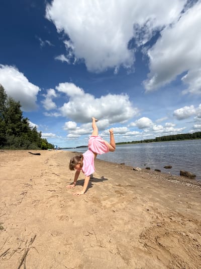The image captures a vibrant outdoor scene on a sunny day at a sandy beach in Rusanovo, Russia. A young girl, dressed in a pink t-shirt and pink shorts, is depicted in dynamic motion, seemingly performing a cartwheel or an acrobatic pose. Her hands are placed on the light brown sand, and one leg is extended high into the air, showcasing her flexibility and active play. Her head is angled downwards, partially obscuring her face as she focuses on her movement.
The setting features a wide expanse of light brown sand, with visible textures and a few scattered pieces of dried vegetation. To the right of the girl, a calm body of water stretches into the distance, reflecting the bright sky. The opposite shore is lined with a dense forest of green trees, suggesting a natural and serene environment. Small rocks and pebbles are visible along the water's edge on the sandy bank.
Above, the sky is a striking deep blue, adorned with numerous fluffy white cumulus clouds, indicating clear and pleasant weather conditions. The bright daylight illuminates the scene, enhancing the cheerful and energetic atmosphere. The image conveys a sense of playful freedom and joy, capturing a moment of active outdoor recreation. No visible text is present in the image.