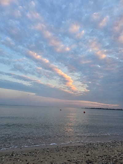 This image depicts a serene beach scene at sunrise or sunset. The sky is filled with soft, wispy clouds tinged with pink and orange hues, suggesting the golden hour. Below the sky, a calm body of water reflects the colors of the sky, with a gentle wave lapping at the shore. The beach itself is composed of sand and small pebbles. In the distance, a faint silhouette of a cityscape can be seen along the horizon. Two red buoys are visible in the water, one closer and one farther away. The overall mood of the image is peaceful and tranquil. The orientation of the image is vertical, as if the photographer was standing and capturing the scene at a slight upward angle.