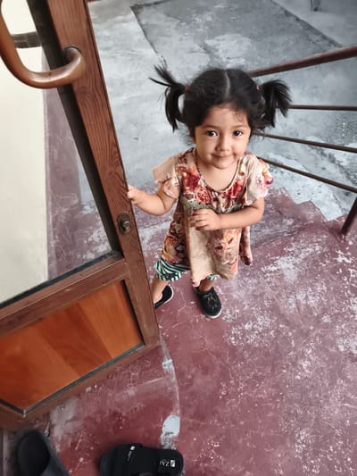 A young girl with dark hair in two pigtails stands on a set of reddish-brown stairs. She is wearing a light-colored, short-sleeved dress with a floral print of pink, red, and orange flowers. Beneath the dress, she appears to be wearing striped shorts, visible at the hem. Her feet are clad in black slip-on shoes with small decorative elements.
The girl is positioned near a wooden door frame with a curved, brass-colored handle and a keyhole. Her right hand is lightly touching the door frame. She is looking directly at the camera with a gentle smile, conveying a sense of innocence and curiosity.
The setting appears to be outdoors, possibly a courtyard or entrance to a building, given the stairs and the concrete or stone ground. The stairs are worn and have a textured, somewhat mottled appearance. Metal railings are visible behind the girl on the right side of the frame. The overall lighting suggests it could be daytime.
The image was taken in Buz, Uzbekistan. No visible text is present in the image. The mood is casual and candid, capturing a moment with a child.
