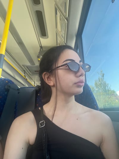 A young woman with dark hair and sunglasses rests her head back against a blue bus seat, looking upwards. The bus has a sunroof, allowing bright daylight to illuminate the scene. Trees and a blue sky are visible through the sunroof. The woman is wearing a black top with a strap across her shoulder. The interior of the bus is visible, with yellow poles and gray paneling. The image appears to be taken during the daytime, with clear weather.