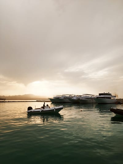 The image depicts a harbor scene in Ras Abu Soma, Egypt, at dusk or dawn. In the foreground, a white inflatable boat with dark trim is occupied by two people; one appears to be steering the vessel. Several large yachts are docked in the background, appearing uniform in color. A landmass is visible in the distance, with faint outlines of mountains under a cloudy sky. The water has ripples.