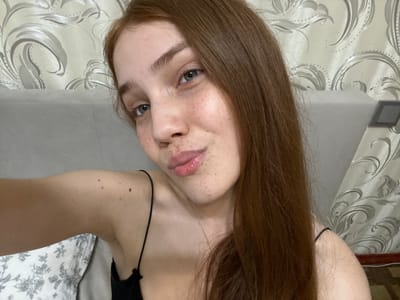 A young woman with long, auburn hair and freckles on her face is taking a selfie indoors. She is wearing a black spaghetti strap top. Her left arm is visible, and she has several moles on her shoulder and upper arm. The background appears to be a room with patterned wallpaper and a headboard of a bed. The lighting is soft and natural. There is no indication of the specific location within Naberezhnye Chelny, Russia, other than it being an indoor setting.