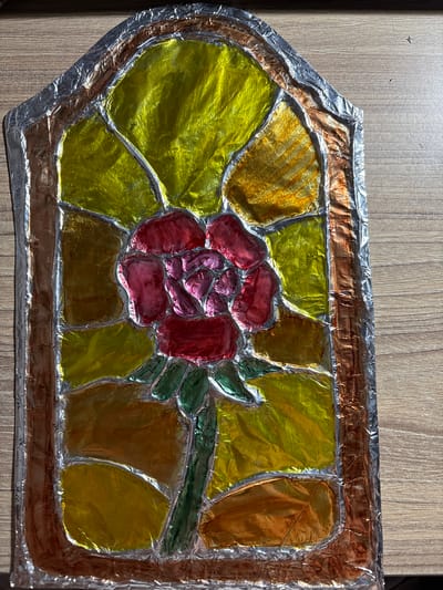 The image displays a homemade stained-glass-style artwork of a rose, created from colorful metallic foil. The artwork is framed with what appears to be brown and silver foil, mimicking leaded glass. The rose is depicted in shades of red and pink, with green foil for the stem and leaves. The background is a patchwork of yellow and orange foil, suggesting petals or light. The entire piece is laid on a wooden surface, likely a table, with a visible wood grain pattern. The lighting casts shadows, highlighting the textured, crinkled surface of the foil. There are no people or animals present in the image. The artwork is positioned on the wooden surface, and no specific location within Panagyurishte, Bulgaria, is indicated or inferable from the image itself.