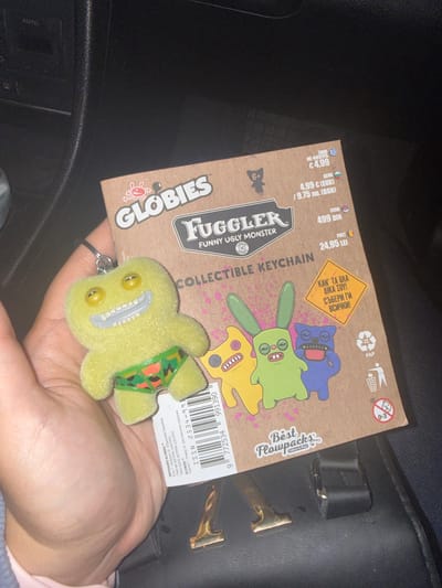 The image shows a hand holding a Globies "Fuggler" collectible keychain. The keychain is yellow and depicts a monster with a green and orange patterned diaper. The keychain is attached to a metal keyring. The packaging for the keychain is brown cardboard. The packaging states "Globies Funny Ugly Monster Collectible Keychain". The packaging also includes pricing in Euro (€4.99), Bulgarian Lev (9.75 лв.), Serbian Dinar (499 DIN) and Romanian Leu (24.95 LEI). The packaging has a warning label for children aged 0-3. The background includes part of a car interior, and a black leather purse. The location cannot be determined as Okop, Bulgaria from the image.