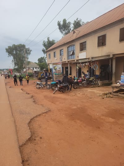 The image depicts a street scene in Bokkos, Nigeria. A two-story building with a beige facade and a brown corrugated roof is on the left. The building has several windows, some of which appear to be boarded up. The ground floor of the building appears to be a shop or business, with items displayed outside, including colorful rings that resemble tires. Several motorcycles are parked in front of the building.

On the right side of the image is a dirt road. People are seen walking and riding motorcycles on the road. Some individuals are wearing dark clothing, while others have on brighter colors. There are trees visible in the background, and power lines stretch across the sky. The sky is overcast, suggesting it might be a cloudy day.

In the foreground of the dirt road, several motorcycles are lined up, with riders on some of them. The ground is a reddish-brown color, characteristic of laterite soil common in the region.

Visible text includes a sign on the building that appears to be an advertisement, with images of power tools and text that is partially legible, reading "BASED KORON COMPUTU [BBC]". Another sign, red in color, is visible further down the road.

The scene suggests a typical day in a Nigerian town, with commerce, transportation via motorcycles, and people going about their daily activities. The time of day is difficult to ascertain precisely due to the overcast sky, but it appears to be daytime. There are no obvious cues for emotions, and the overall impression is one of everyday life.