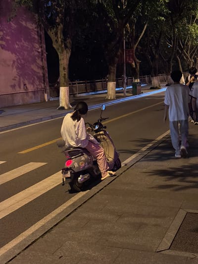 A woman is riding a moped on a street in Nan'an District, China. The rider wears a white shirt and pink pants. The moped has a backpack attached and is parked on a crosswalk. Another person walks along the sidewalk, facing away from the moped. Trees line the street.