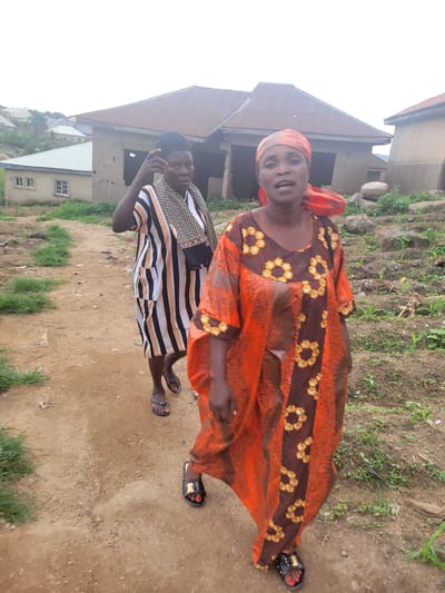 In Madalla, Nigeria, two women walk on a dirt path. The woman in the foreground wears an orange and brown patterned dress and a headscarf. The woman behind her is dressed in a striped tunic and wears a head covering. The background shows several buildings with corrugated metal roofs, some appearing unfinished, and vegetation. The scene depicts a residential area with a mix of constructed and developing structures.