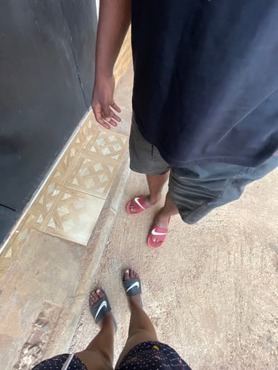 The image depicts a view looking down at two individuals standing near an edge, likely a sidewalk or curb. The person on the left is wearing a dark-colored top, dark shorts, and red Nike slides. Their arm is visible, with an open hand gesture. The person on the right is wearing a patterned dark garment, with black Nike slides on their feet. The ground is mostly a light brown concrete texture. There's an area of decorative tiles at the edge. The location is Kurunduma, Nigeria, as indicated in the prompt.
