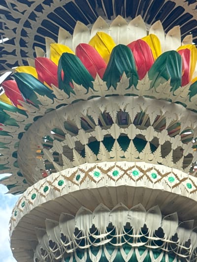This is a close-up, low-angle, vertical shot of the top of a traditional Balinese *penjor*, a ceremonial bamboo pole, in Tegallalang, Indonesia. The *penjor* is intricately decorated with woven palm leaves and colorful fabrics in red, green, and yellow, arranged in a tiered, fan-like structure. The outer layers are made of white, leaf-shaped woven elements with circular green embellishments. The sky is visible in the upper left corner.