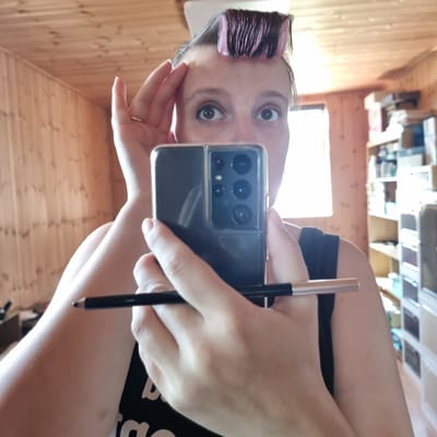 A woman is reflected in a mirror, holding a smartphone in her right hand and a makeup pencil in her left. She is wearing hair rollers on the top of her head and a black tank top. The background shows a wooden paneled wall and shelves filled with various items. The scene appears to be indoors, possibly in a home. There is no information or visual cues to suggest the location is Dunnae-myeon, South Korea.