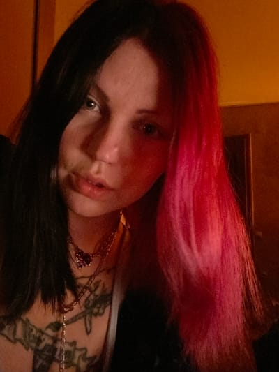 A close-up, rotated view of a fair-skinned person with black and pink hair. The hair is parted, with pink hair at the top of the frame and black hair near the bottom. The person has light-colored eyes and is wearing a gold-colored necklace with a red pendant. A tattoo is visible on their chest. The background is a blurred mix of dark and light shades of brown. The location is not evident in the image, and there is no visual reference to indicate the city of Kunje, Montenegro.