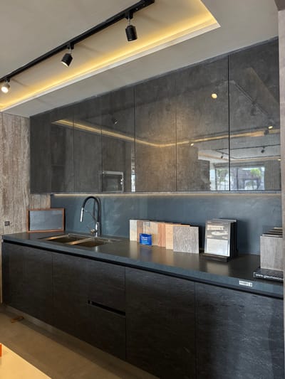The image displays a modern kitchen or kitchen showroom installation located in Delegación Josefa Vergara y Hernández, Mexico. The scene is devoid of people.

The environment features a dark, contemporary aesthetic. Overhead, a light-colored ceiling incorporates a black track lighting system with several adjustable spotlights directed downwards. A recessed linear light channel running parallel to the upper cabinets emits a warm, diffused glow.

Below the ceiling, a row of dark grey, highly reflective upper cabinets with a subtle texture reflects the overhead lighting and elements of the surrounding space, including a partial view of an exterior or an adjacent room through what appears to be a window or doorway. A linear LED light strip illuminates the dark, textured backsplash situated between the upper cabinets and the countertop.

The countertop is a matte dark grey or black material, likely stone. It integrates a double-basin stainless steel sink equipped with a black, flexible gooseneck faucet. Beneath the countertop, the base cabinets are dark grey or black with a textured finish, resembling slate, featuring integrated horizontal handles for drawers and doors.

On the right side of the countertop, multiple material samples are displayed: a series of upright rectangular tiles or slabs in various shades of light wood, beige, and grey are arranged sequentially. Adjacent to these, a black stand holds several design brochures or booklets, and a larger vertical sample display is partially visible, with "www.giecmexico.co" printed on its base. To the far left on the countertop, a small, dark-framed board rests against the backsplash. The floor is a light-colored, possibly concrete or large tile, surface. No actions or interactions are depicted.