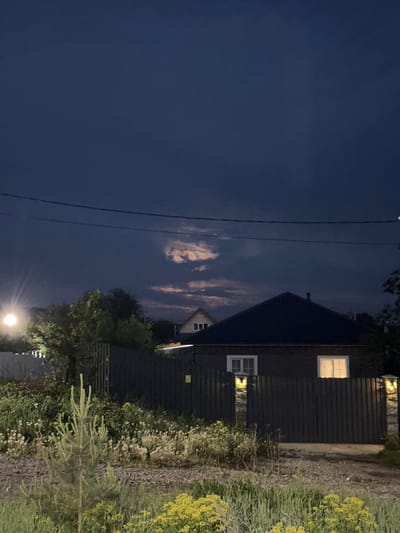 The image depicts a night scene in Горный, Russia. A wooden house is visible with lit windows, adjacent to a gravel path bordered by overgrown vegetation. In the foreground, power lines stretch across a dark blue sky with patches of clouds. A street lamp illuminates the scene from below.