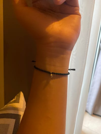 A close-up, low-angle shot shows a person's forearm and wrist wearing a thin, dark braided bracelet. The bracelet has a small, shiny charm dangling from it, possibly a bead or a small metallic object. The skin tone is warm and tanned. The background is dimly lit and out of focus, with a vertical pattern that appears to be a sheer curtain or blind on the right side of the frame, and a textured, patterned cushion on the left. The lighting suggests an indoor setting, likely during the daytime given the presence of natural light filtering through the curtain. There are no visible elements that definitively place this image within Central Jakarta, Indonesia.
