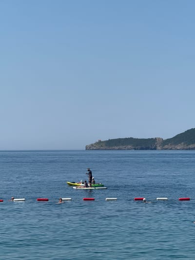 The image depicts a serene coastal scene in Rafailovići, Montenegro, under a clear, bright blue sky. The tranquil, deep blue sea fills the lower half of the frame, with a discernible horizon line separating it from the sky. A line of red and white buoys, marking a swimming area, stretches across the foreground, with several swimmers visible in the water. In the middle ground, a group is engaged in water activities on two inflatable paddleboards and a yellow kayak. One person stands on a paddleboard, appearing to guide the group, while others are seated or lying down on the boards. The background features a lush green, hilly coastline dotted with trees and a small building perched on the highest point of a prominent headland. The overall impression is one of a peaceful summer day at the seaside.