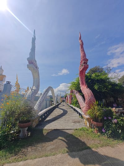 A bridge lined with mythical serpent sculptures under a bright sky. The serpents, one white and one red, rise dramatically from the bridge's edge, forming decorative archways. Lush greenery flanks the bridge, adding a natural element to the ornate structure. Sunlight casts distinct shadows across the pathway, highlighting its curvature and leading the eye toward the distant end. Potted plants enhance the bridge's aesthetics.