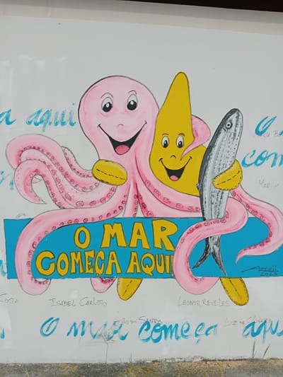 This is a vibrant and cheerful cartoon-style mural, likely painted on an outdoor wall or panel in Figueira da Foz, Portugal. The main subjects are a large, smiling pink octopus and a smaller, happy yellow star, both interacting with a realistic-looking silver fish.
The pink octopus, with its numerous tentacles, playfully holds the fish across its body using two of its limbs, which appear to have yellow cuff-like markings. Its round eyes and wide smile convey a jovial expression. Below the fish, the yellow star also sports a wide smile and large eyes, seemingly supporting or engaging with the fish. The fish itself is depicted with notable detail, including scales and a textured appearance, contrasting with the smooth, cartoonish style of the octopus and star.
A prominent vertical blue banner runs through the right side of the artwork, featuring bold yellow text that reads, "O MAR COMEÇA AQUI" (The Sea Starts Here), clearly indicating a marine theme and connection to the ocean. Around the central illustration, numerous signatures and names are written in blue and black, suggesting multiple contributors or dedications to the artwork. Visible names include "MARIA BE", "MARTIM", "LEONOR REVELES", "LUIZ DE OLIVEIRA", "JOÃO DOS SANTOS", "ISABEL CARLOTO", and "COSTA". A date, "2025", is also visible near the top right of the blue banner. The overall impression is one of a community art project, possibly for children or celebrating a local connection to the sea. The bright colors and playful expressions create a joyful and welcoming atmosphere.