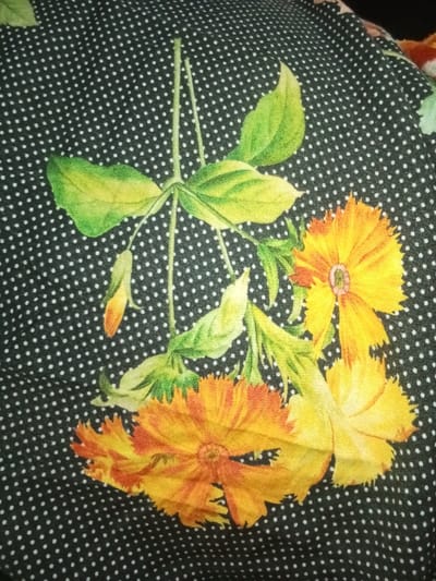 The image displays a close-up of a patterned fabric. The background of the fabric is dark, likely black or deep grey, adorned with a uniform pattern of small, white polka dots. Superimposed on this dotted background is a floral print featuring bright yellow and orange flowers with green foliage. There are at least three prominent flowers, characterized by multiple petals and visible central structures, alongside several green leaves and what appear to be flower buds. The leaves exhibit variations in green hue, with some areas appearing yellowish-green. The fabric itself shows slight wrinkles and folds. No people, discernible actions, or environmental context beyond the fabric are visible. The specific location within Umsobomvu Local Municipality, South Africa, cannot be determined from the content of this image.