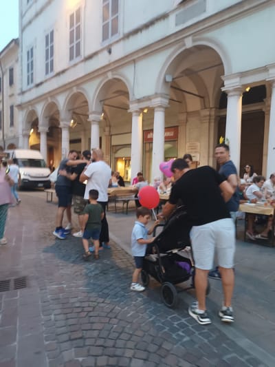 A group of people are gathered on a cobblestone street in front of an arcade of arches. The architecture suggests a European town, possibly Italy, given the context of Viadana. In the background, there is a white van. Several tables with people seated at them are visible, indicating a gathering or outdoor dining event. Two red and pink balloons are also visible, suggesting a celebration or festive occasion. A man is pushing a stroller with a young child. The lighting suggests it is dusk or early evening. The atmosphere appears relaxed and social. There is some visible text on a sign that appears to read "alt" and "neu" but the rest is illegible.