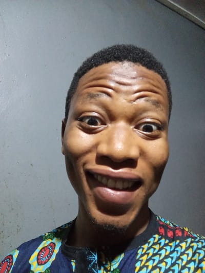 A head-and-shoulders shot shows a young man with dark skin and short, dark hair against a gray wall. He is smiling widely, showing his teeth, and his eyes are wide. He is wearing a colorful shirt with a blue background and patterns featuring red, yellow, and green elements. No elements suggesting a location in Oshogbo, Nigeria, are visible.