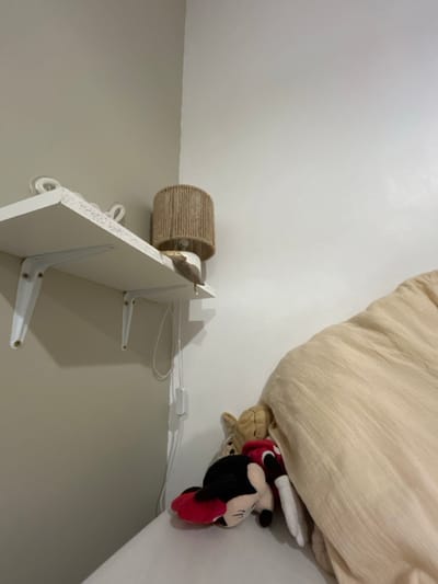 The image shows a corner of a room with a bed, a bedside lamp, and a white shelf. The bed has light beige bedding. Under the edge of the bedding are two plush toys, a brown one and a Mickey Mouse toy. The bedside lamp has a woven cylindrical shade. The white shelf is mounted on the wall with white brackets. A white cable runs along the wall near the shelf and the bed. The walls are painted in light grey and white.