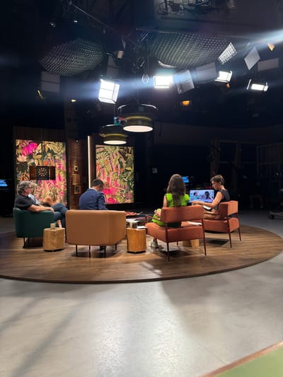 The image depicts a television studio set, likely located in the city of Sant Joan Despí, Spain, known for its media production facilities. The set features a circular wooden platform on a concrete floor, with several seating arrangements. In the background, two large panels display vibrant tropical foliage with pink and green hues. A dartboard is mounted on the left panel.
Five individuals are present:
* On the left, a man with graying hair is seated in a dark green armchair, facing inward. Next to him, a woman with blonde hair is also seated in the armchair, her body angled towards the center.
* In the middle, a man in a blue suit is seated in a tan armchair, facing away from the camera.
* On the right, two women are seated in tan armchairs. One woman, facing away from the camera, wears a bright green outfit. The other woman, facing towards a monitor, wears a black sleeveless top and a light brown skirt.
Several objects are visible:
* Two cylindrical wooden stools are placed near the armchairs.
* A low-profile coffee table with some items on it is situated in the center of the seating arrangement.
* A large monitor is positioned on a stand behind the two women on the right.
* Studio lighting equipment, including large softboxes and overhead lights, is visible in the dark ceiling.
* Camera equipment and other production elements are present in the background, suggesting an active filming environment.
The overall scene suggests a talk show or panel discussion being recorded in a professional studio setting.