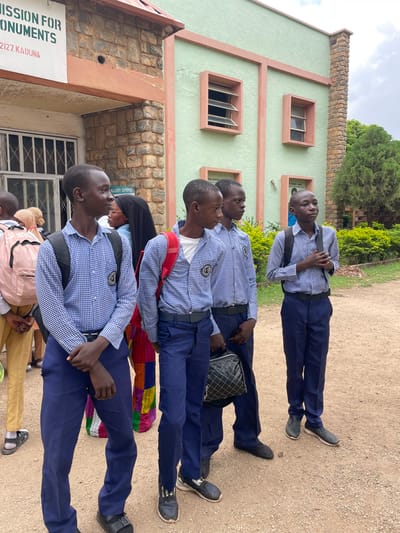 The image depicts a group of young people, likely students, standing outside a light green building with a stone facade. The building has several windows and appears to be a public institution, possibly a museum or gallery, as indicated by a sign that reads "ISSION FOR MONUMENTS" and "2127 KADUNA".
The subjects are all dressed in uniforms consisting of blue and white checkered shirts and dark blue trousers. Most are wearing backpacks. Some of the individuals are looking up and to the side, suggesting they are waiting or observing something. The overall mood seems neutral and organized.
The scene is set outdoors on a dirt ground with scattered gravel, under what appears to be a cloudy sky. There are trees and bushes in the background, indicating a location with some greenery. The lighting suggests it is daytime. The orientation of the image is on its side.