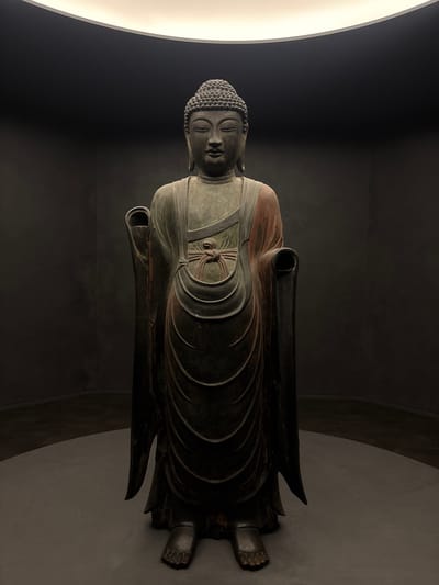 A full-body, front-facing bronze statue of Buddha stands in a dimly lit, circular room. The statue is positioned on a circular platform, with a subtle spotlight illuminating it from above. The background consists of dark, textured walls that curve away from the statue. The Buddha statue is depicted in a standing pose, with its right hand raised and palm facing outward, and its left hand lowered. It wears traditional monastic robes, with one shoulder exposed and draped with a reddish-brown fabric. The statue has a serene expression with closed eyes and a coiled ushnisha on its head. The lighting casts long shadows, emphasizing the statue's form and the texture of the robes. The overall atmosphere is solemn and contemplative. This statue is located at Bulguksa Temple in Gyeongju-si, South Korea.