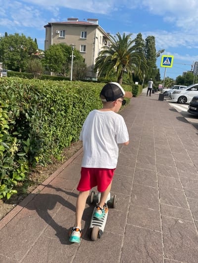 A young boy is riding a scooter on a paved sidewalk in Gagra, Abkhazia, on a sunny day. The boy, wearing a white t-shirt, red shorts, a baseball cap, and colorful sneakers, is moving away from the camera. A neatly trimmed green hedge runs along the left side of the sidewalk. Behind the hedge, a light-colored building with a red roof is visible, with trees and palm trees surrounding it. Across the street, several cars are parked along the curb, and a pedestrian crossing sign is posted. The sky is blue with scattered white clouds.