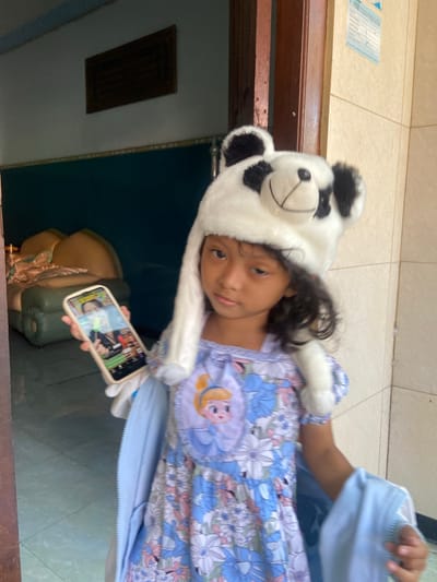 A young girl wearing a panda hat and a blue floral dress stands in what appears to be the entrance of a building. She is holding a smartphone in her left hand, displaying a video call. The background suggests an indoor setting with a doorway, tiled walls, and a couch visible in a darker room behind her. The location is identified as Gelam, Indonesia.